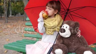 小孩坐在靠近一只红色伞下的<strong>玩具熊</strong>的木凳上，在公园里吃<strong>大</strong>红
